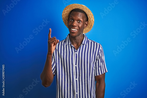 African american man wearing striped shirt and summer hat over isolated blue background pointing finger up with successful idea. Exited and happy. Number one.