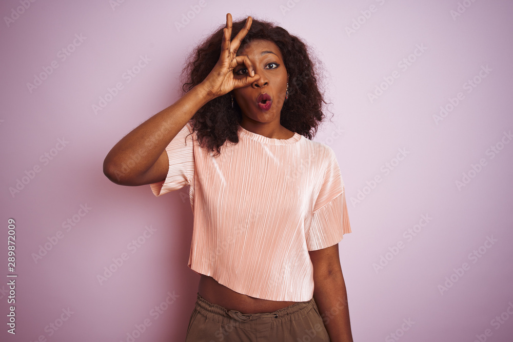 Young african american woman wearing t-shirt standing over isolated pink background doing ok gesture shocked with surprised face, eye looking through fingers. Unbelieving expression. Young african american woman wearing t-shirt standing over isolated pink background doing ok gesture shocked with surprised face, eye looking through fingers. Unbelieving expression.