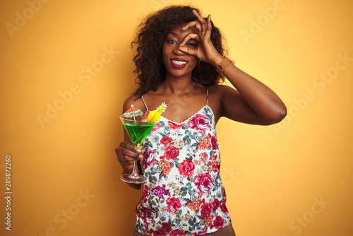 Young african american woman drinking cocktail standing over isolated yellow background with happy face smiling doing ok sign with hand on eye looking through fingers