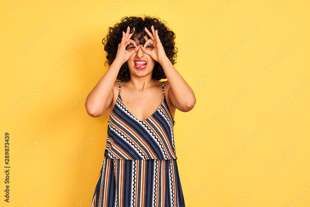 Arab woman with curly hair wearing striped colorful dress over isolated yellow background doing ok gesture like binoculars sticking tongue out, eyes looking through fingers. Crazy expression. Arab woman with curly hair wearing striped colorful dress over isolated yellow background doing ok gesture like binoculars sticking tongue out, eyes looking through fingers. Crazy expression.