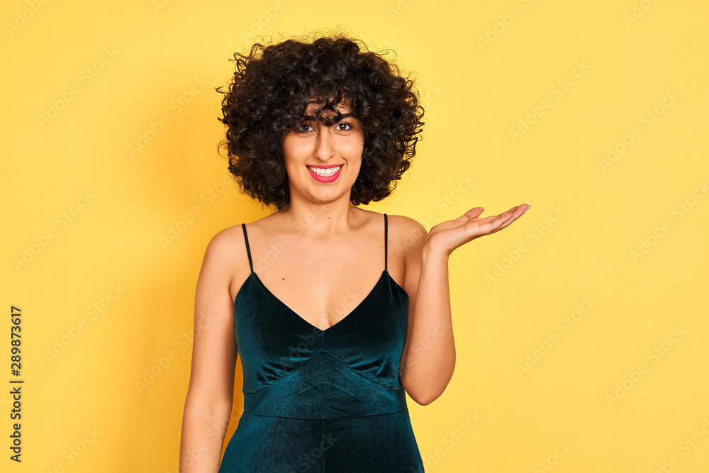 Young arab woman with curly hair wearing elegant dress over isolated yellow background smiling cheerful presenting and pointing with palm of hand looking at the camera. Young arab woman with curly hair wearing elegant dress over isolated yellow background smiling cheerful presenting and pointing with palm of hand looking at the camera.