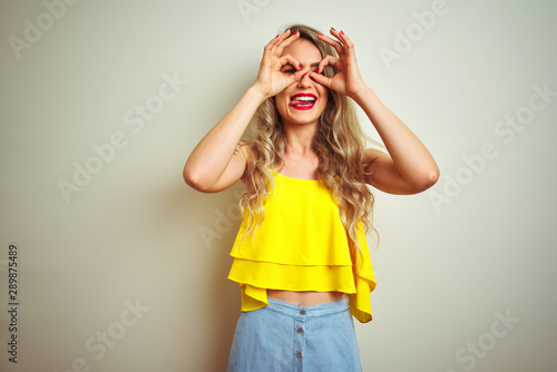 Young beautiful woman wearing yellow t-shirt standing over white isolated background doing ok gesture like binoculars sticking tongue out, eyes looking through fingers. Crazy expression.