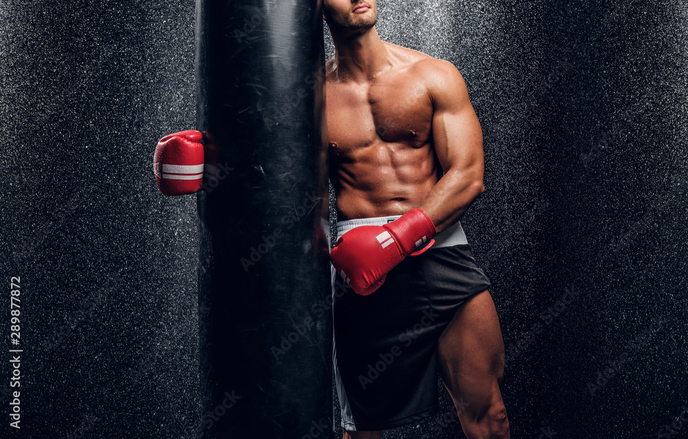 Young muscular man in red boxer gloves is posing with punching bag at dark room with water splashes on the wall. Young muscular man in red boxer gloves is posing with punching bag at dark room with water splashes on the wall.