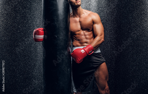 Young muscular man in red boxer gloves is posing with punching bag at dark room with water splashes on the wall. © Fxquadro