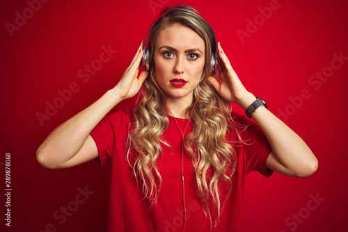 Young beautiful woman wearing headphones over red isolated background with serious expression on face. Simple and natural looking at the camera.