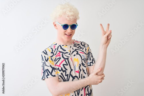 Young albino blond man wearing colorful t-shirt and sunglasses over isolated red background with a big smile on face, pointing with hand and finger to the side looking at the camera.