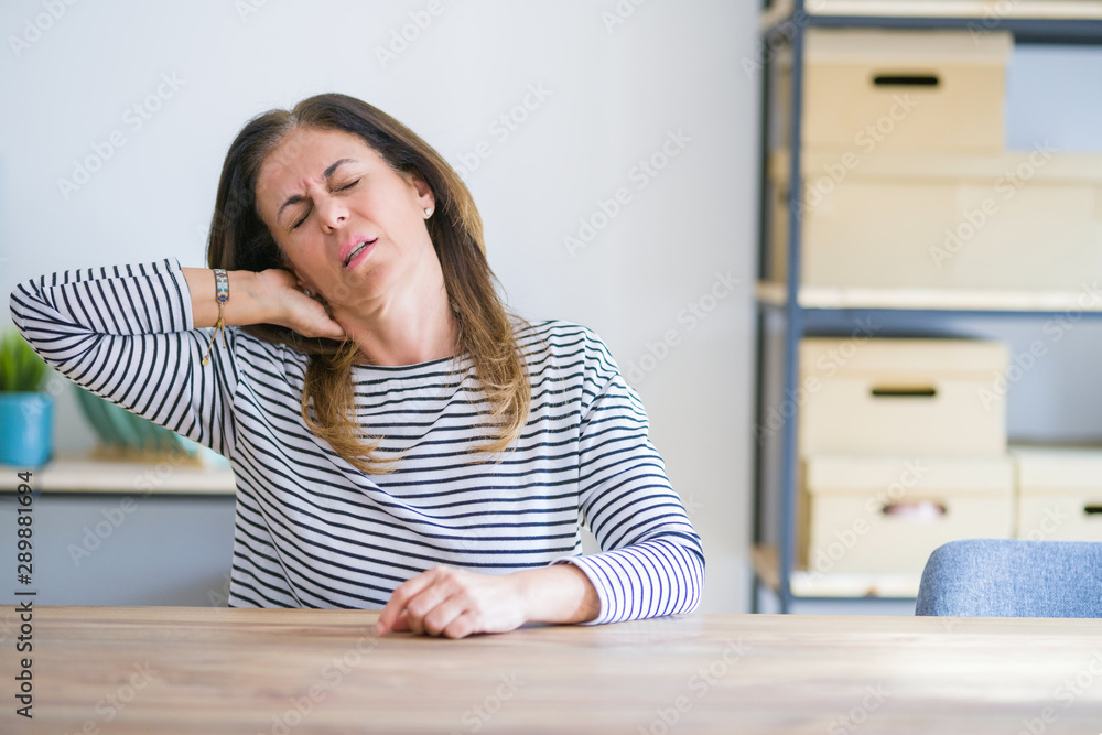 Middle age senior woman sitting at the table at home Suffering of neck ache injury, touching neck with hand, muscular pain Middle age senior woman sitting at the table at home Suffering of neck ache injury, touching neck with hand, muscular pain