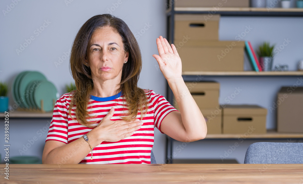 Middle age senior woman sitting at the table at home Swearing with hand on chest and open palm, making a loyalty promise oath Middle age senior woman sitting at the table at home Swearing with hand on chest and open palm, making a loyalty promise oath