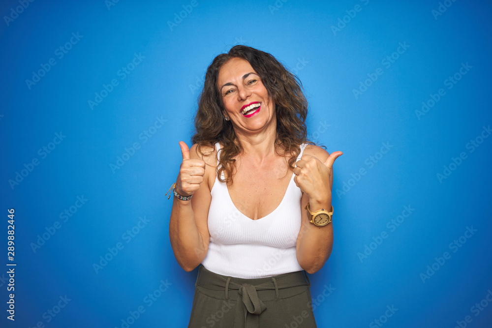 Fototapeta premium Middle age senior woman with curly hair standing over blue isolated background success sign doing positive gesture with hand, thumbs up smiling and happy. Cheerful expression and winner gesture.