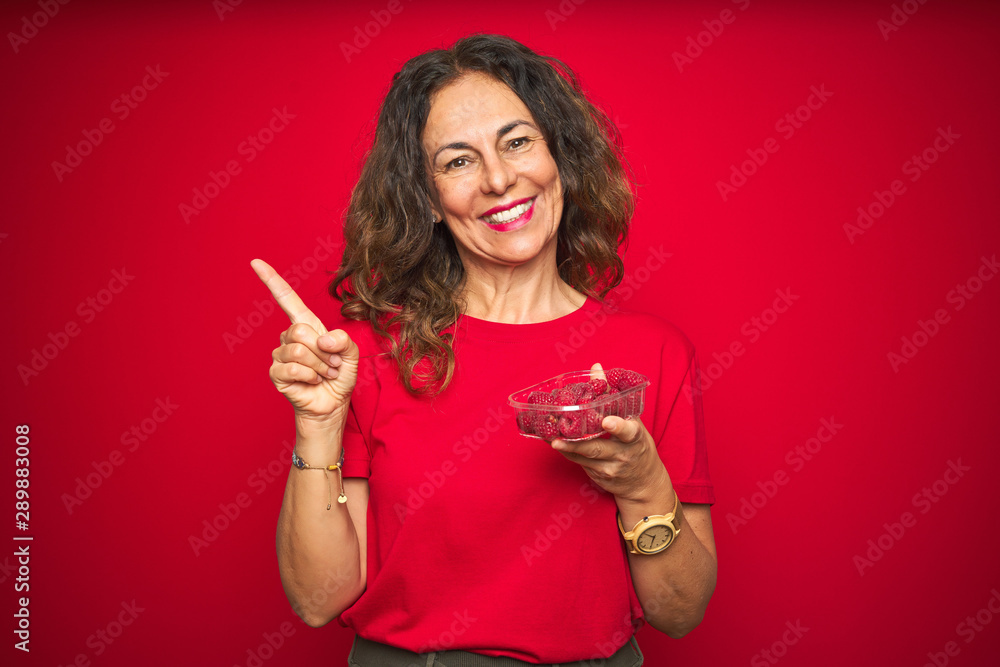 Naklejka premium Middle age senior woman eating raspberries over red isolated background very happy pointing with hand and finger to the side