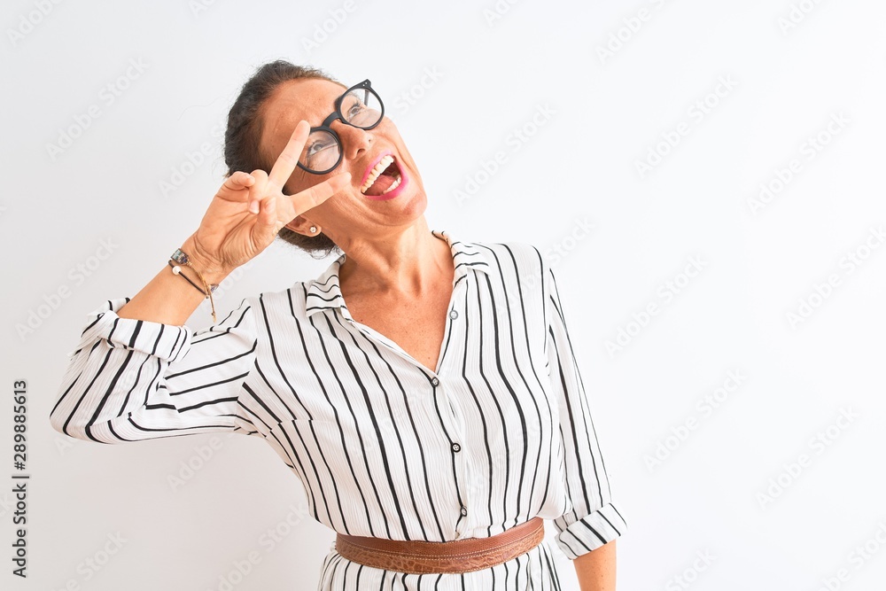 Middle age businesswoman wearing striped dress and glasses over isolated white background Doing peace symbol with fingers over face, smiling cheerful showing victory Middle age businesswoman wearing striped dress and glasses over isolated white background Doing peace symbol with fingers over face, smiling cheerful showing victory