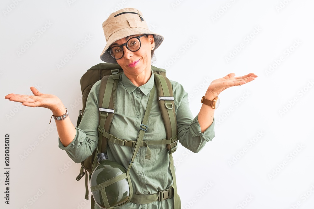 Middle age hiker woman wearing backpack canteen hat glasses over isolated white background clueless and confused expression with arms and hands raised. Doubt concept. Middle age hiker woman wearing backpack canteen hat glasses over isolated white background clueless and confused expression with arms and hands raised. Doubt concept.