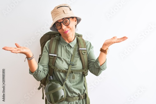 Middle age hiker woman wearing backpack canteen hat glasses over isolated white background clueless and confused expression with arms and hands raised. Doubt concept.