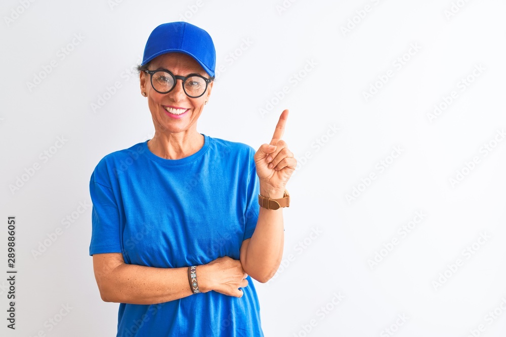 Senior deliverywoman wearing cap and glasses standing over isolated white background with a big smile on face, pointing with hand and finger to the side looking at the camera.