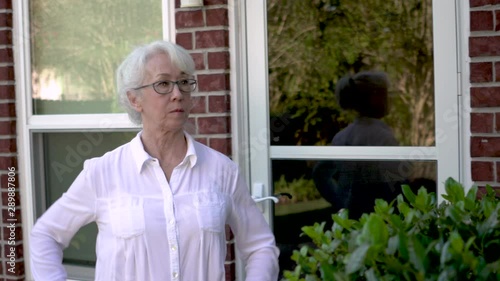 A mature woman walks out of the backdoor of her home looks around briefly then walks past the camera.