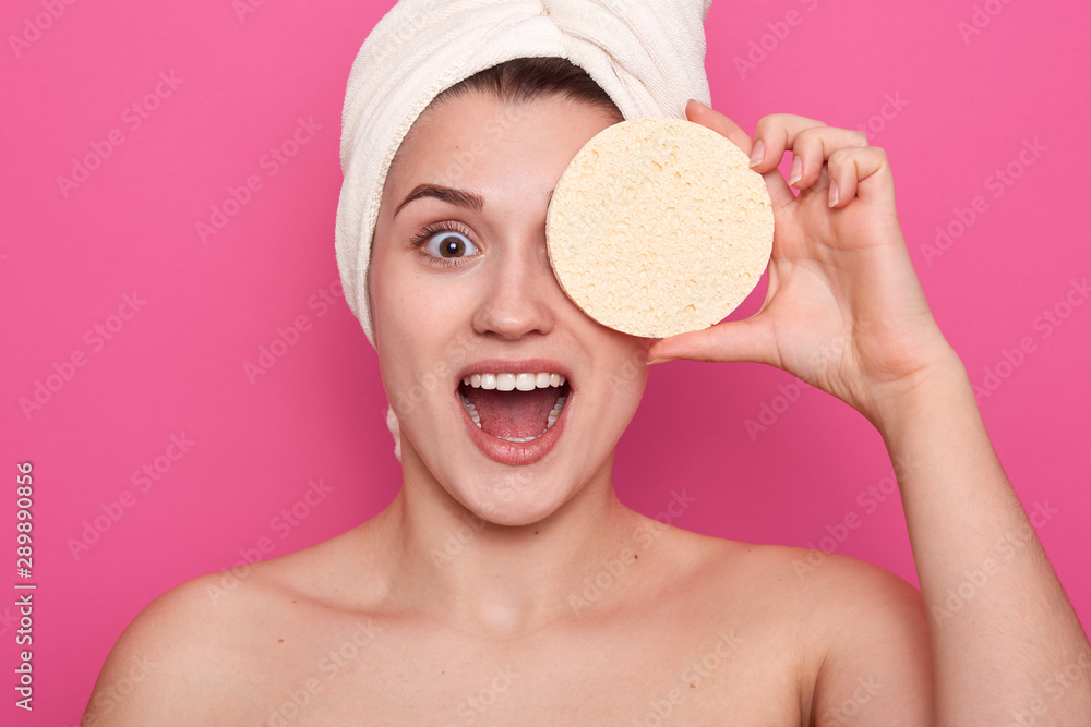 Φωτογραφία Studio shot of smiling woman with cosmetic sponge covering one eye,wrapped with white towel, female having perfect skin, standing with widely open mouth, posing over pink background