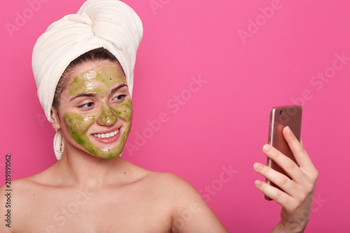 Beautiful female having green mask on face, wrapped white towel, poses half naked, taking selfie on smartphone in bathroom, posing isolated over pink background. Cosmetology and beauty concept. photo