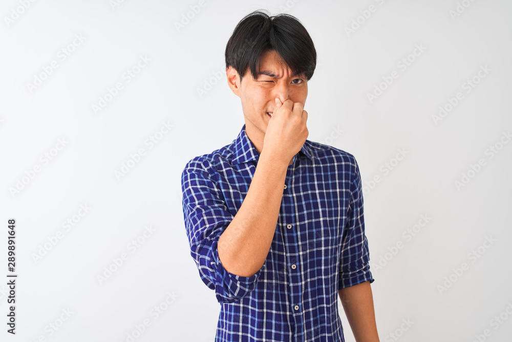 Young chinese man wearing casual blue shirt standing over isolated white background smelling something stinky and disgusting, intolerable smell, holding breath with fingers on nose. Bad smells Young chinese man wearing casual blue shirt standing over isolated white background smelling something stinky and disgusting, intolerable smell, holding breath with fingers on nose. Bad smells
