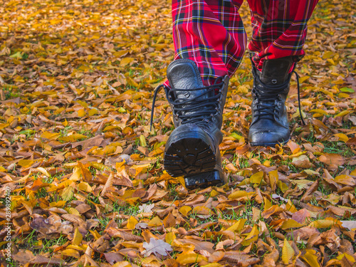 Feet in leather boots with untied shoelaces walking on fall leaves. Outdoor, season clothes, lifestyle, free style with Scottish checked fabric red tartan.