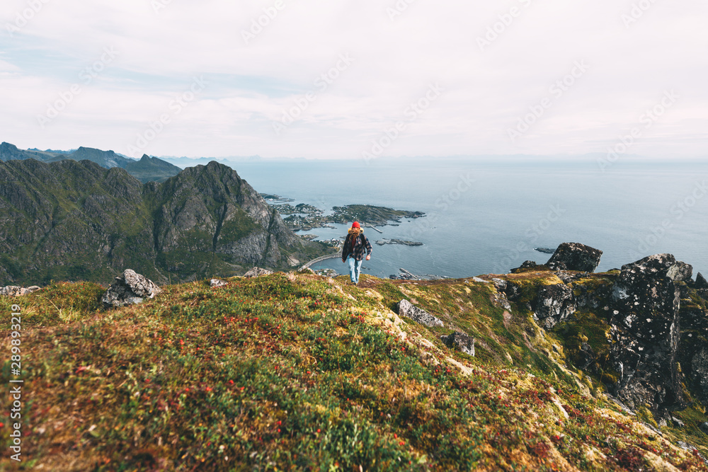 Traveler man wearing backpack and red hat climb on high mountains above sea. Professional expeditor standing on the edge cliff rock and looking forward away. Wanderlust
