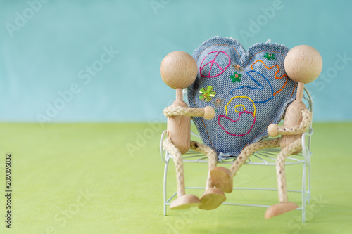Two figurines sitting on a bench, holding a huge sewn heart between them with the embroidered word love
