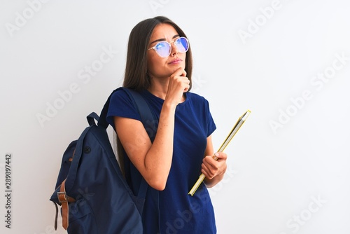 Young student woman wearing backpack glasses holding book over isolated white background serious face thinking about question, very confused idea