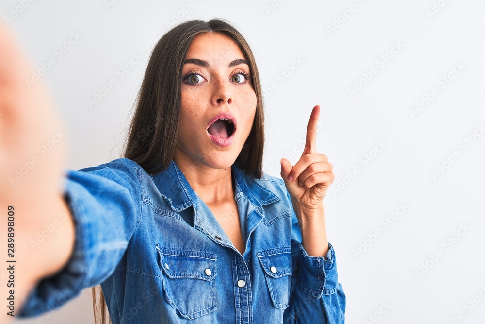Beautiful woman wearing denim shirt make selfie by camera over isolated white background pointing finger up with successful idea. Exited and happy. Number one. Beautiful woman wearing denim shirt make selfie by camera over isolated white background pointing finger up with successful idea. Exited and happy. Number one.
