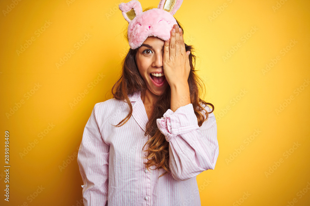 Young woman wearing pajama and sleep mask standing over yellow isolated background covering one eye with hand, confident smile on face and surprise emotion. Young woman wearing pajama and sleep mask standing over yellow isolated background covering one eye with hand, confident smile on face and surprise emotion.
