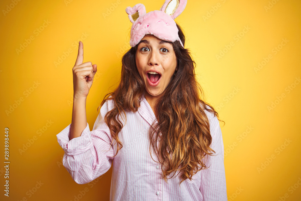 Φωτογραφία Young woman wearing pajama and sleep mask standing over yellow isolated background pointing finger up with successful idea