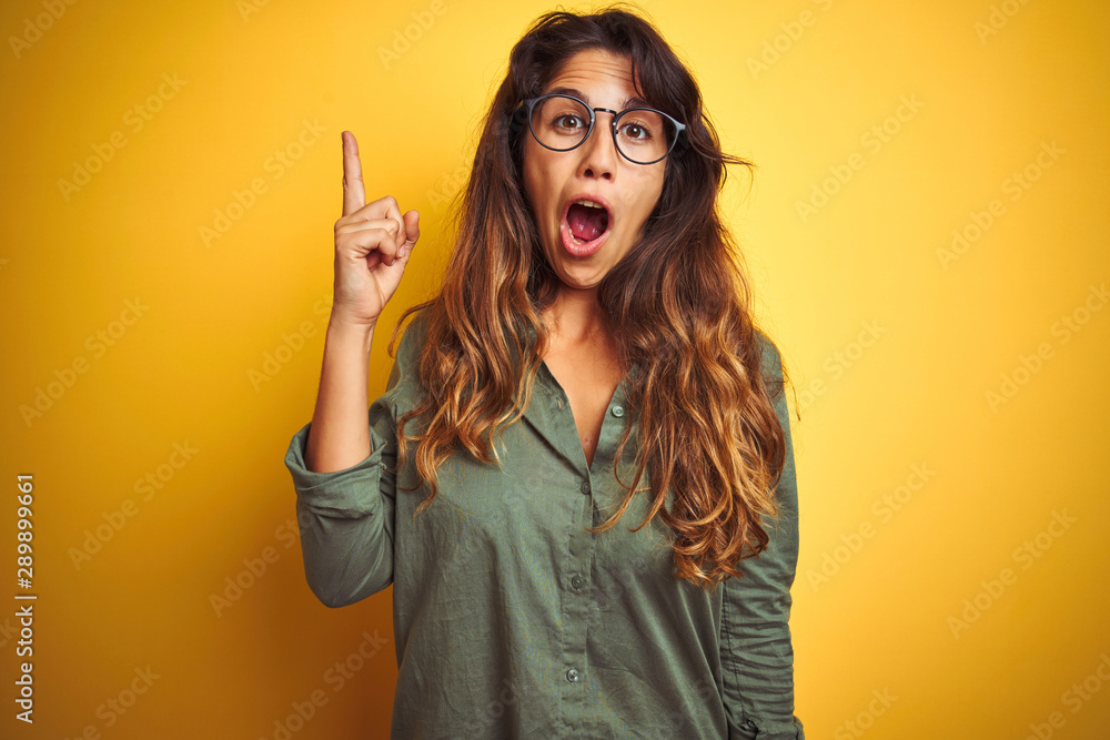 Young beautiful woman wearing green shirt and glasses over yelllow isolated background pointing finger up with successful idea. Exited and happy. Number one. Young beautiful woman wearing green shirt and glasses over yelllow isolated background pointing finger up with successful idea. Exited and happy. Number one.