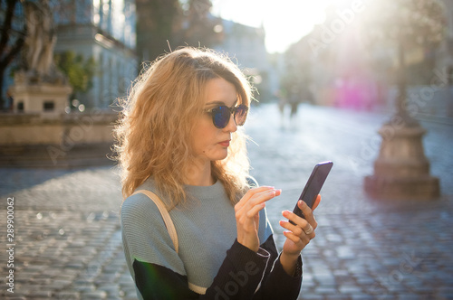 Side view of young woman in blue sunglasses is looking for direction using navigation in her smartphone early in the morning in ancient European city on empty square