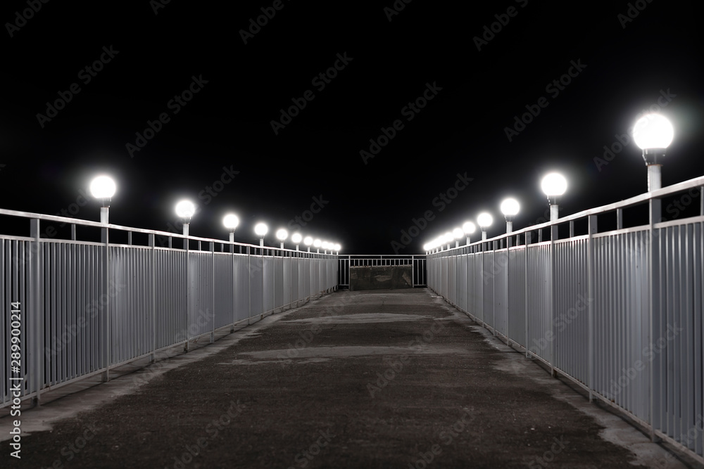 Night sea pier with white lanterns. Round white lights illuminate the track with white iron railings. Walking pedestrian.