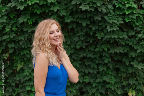 Beautiful young girl smiles in a blue dress, against a background of hedges of wild grapes. Copy space.