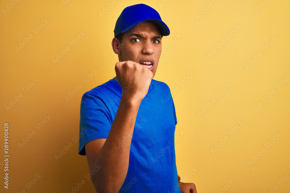 Young handsome arab delivery man standing over isolated yellow background angry and mad raising fist frustrated and furious while shouting with anger. Rage and aggressive concept.