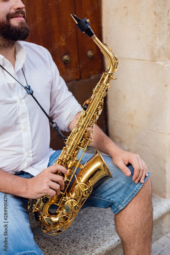 Close up of a man sitting at the door of a building on the street holding his saxophone