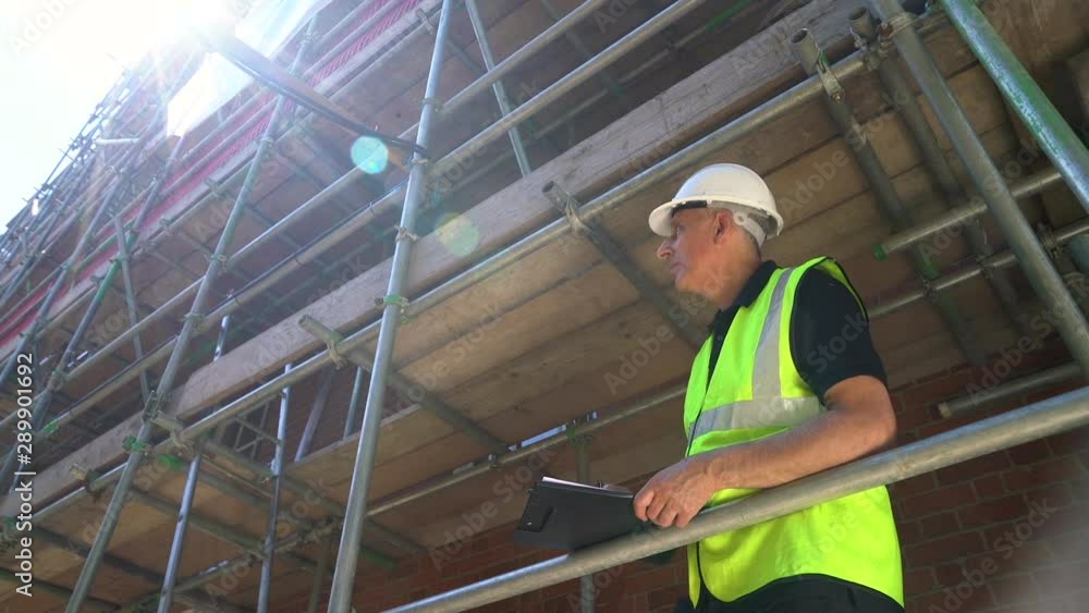 Moving camera clip of male builder foreman, construction worker, surveyor, or architect working on construction building site standing on scaffolding writing on clipboard Moving camera clip of male builder foreman, construction worker, surveyor, or architect working on construction building site standing on scaffolding writing on clipboard