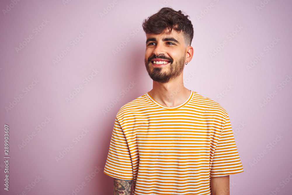 Young man with tattoo wearing yellow striped t-shirt standing over isolated pink background looking away to side with smile on face, natural expression. Laughing confident. Young man with tattoo wearing yellow striped t-shirt standing over isolated pink background looking away to side with smile on face, natural expression. Laughing confident.