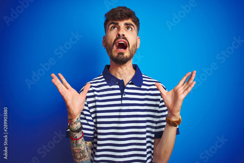 Young man with tattoo wearing striped polo standing over isolated blue background crazy and mad shouting and yelling with aggressive expression and arms raised. Frustration concept.