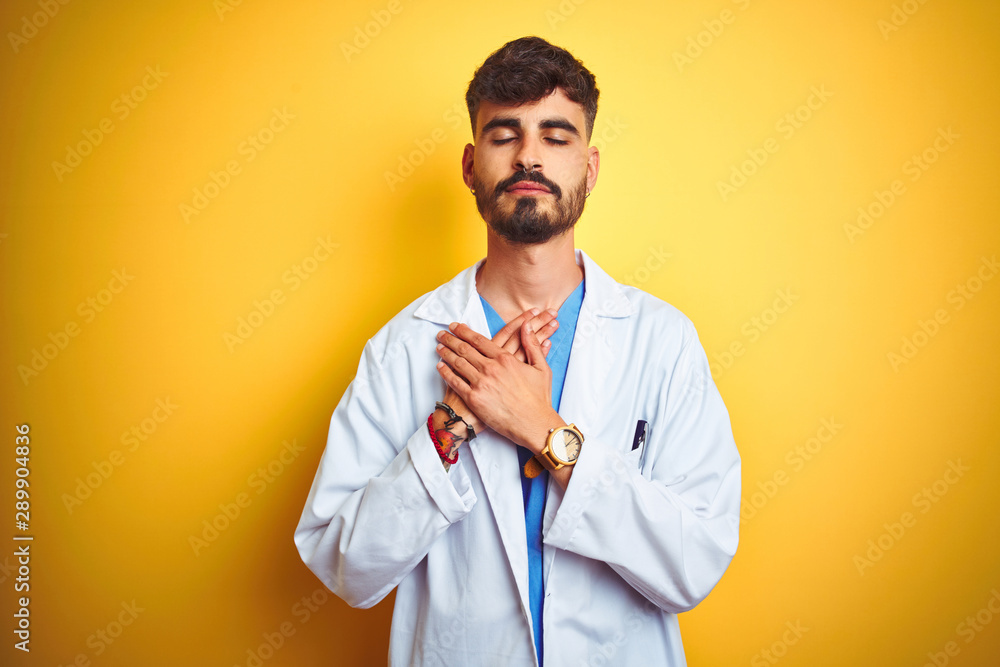 Young doctor man with tattoo standing over isolated yellow background smiling with hands on chest with closed eyes and grateful gesture on face. Health concept.