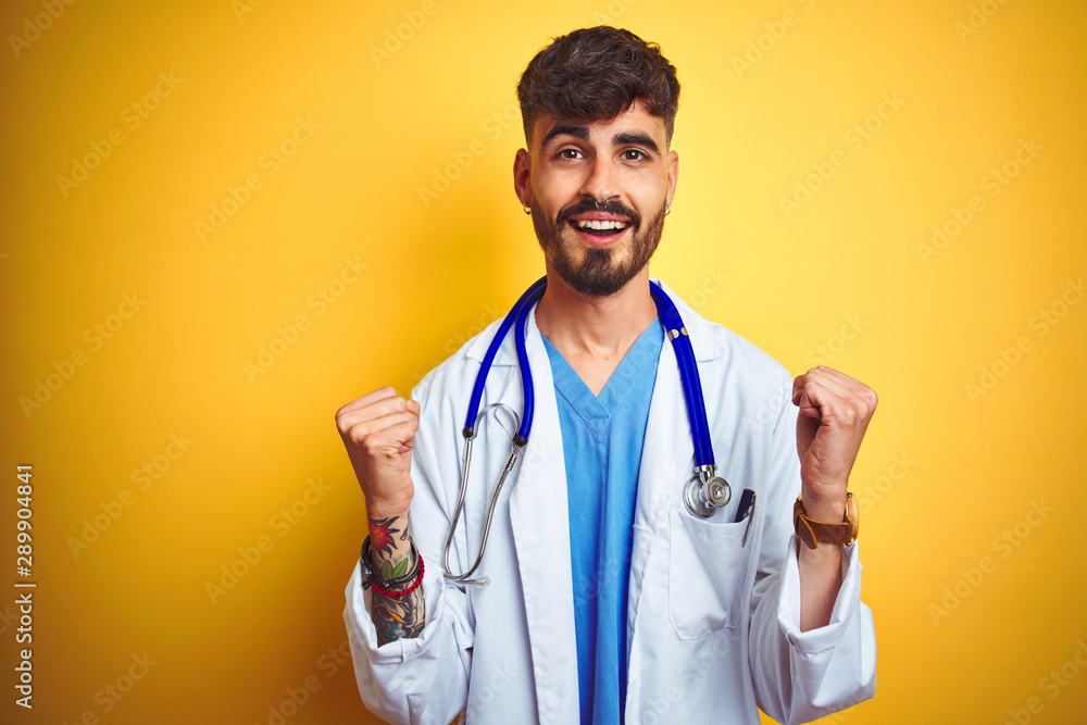 Young doctor man with tattoo wearing stethocope standing over isolated yellow background celebrating surprised and amazed for success with arms raised and open eyes. Winner concept. Young doctor man with tattoo wearing stethocope standing over isolated yellow background celebrating surprised and amazed for success with arms raised and open eyes. Winner concept.