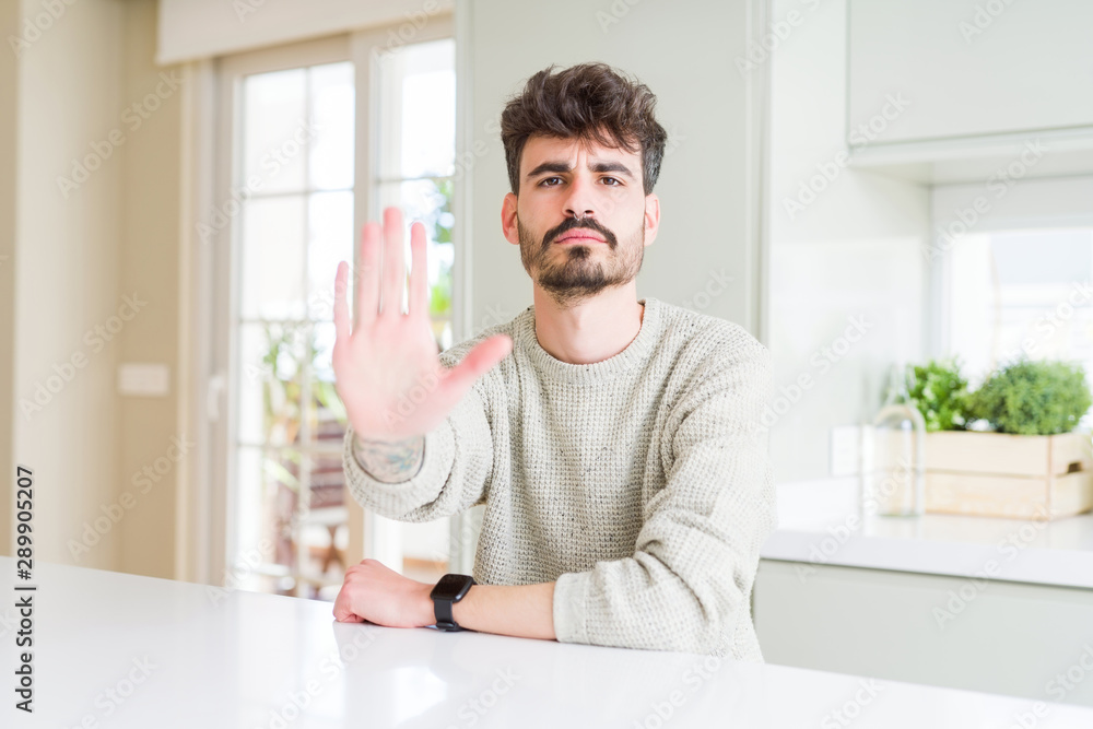 Young man wearing casual sweater sitting on white table doing stop sing with palm of the hand. Warning expression with negative and serious gesture on the face.