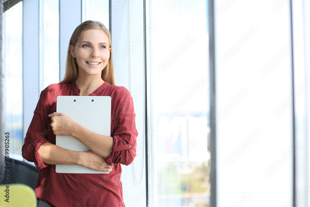 Attractive business woman looking at camera and smiling while standing in the office near the window Attractive business woman looking at camera and smiling while standing in the office near the window