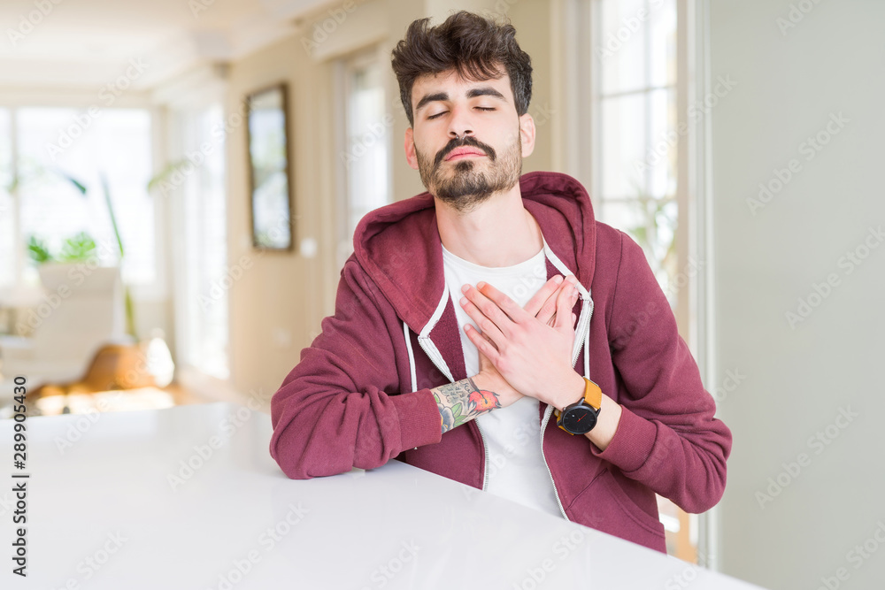 Young man wearing casual sweatshirt sitting on white table smiling with hands on chest with closed eyes and grateful gesture on face. Health concept.