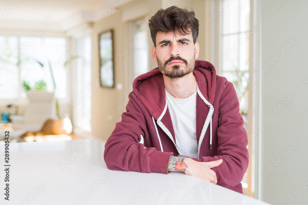 Young man wearing casual sweatshirt sitting on white table skeptic and nervous, disapproving expression on face with crossed arms. Negative person.