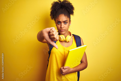 Afro woman using backpack and headphones holding notebook over isolated yellow background with angry face, negative sign showing dislike with thumbs down, rejection concept