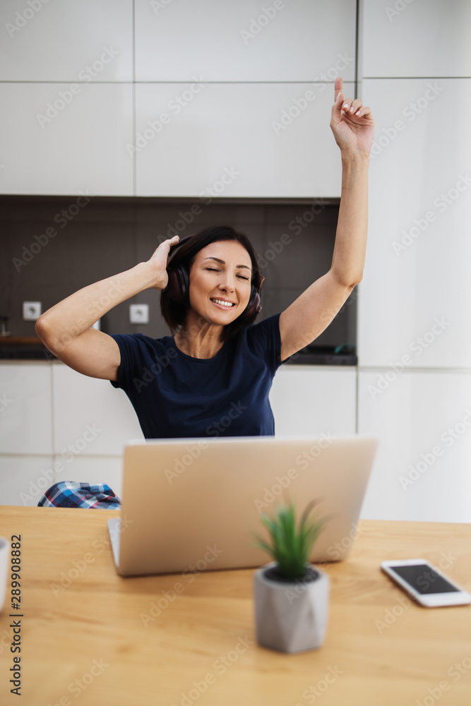 Attractive and happy middle age female freelancer is working at her home. She is using headphones, listening to music and singing. Modern kitchen in background. Freelancing job concept. Attractive and happy middle age female freelancer is working at her home. She is using headphones, listening to music and singing. Modern kitchen in background. Freelancing job concept.