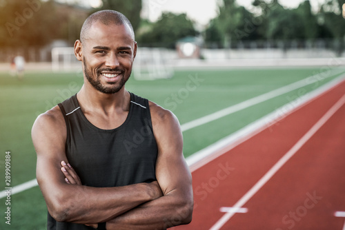 Portrait of cheerful muscular african man standing on a track field outdoors and looking at camera