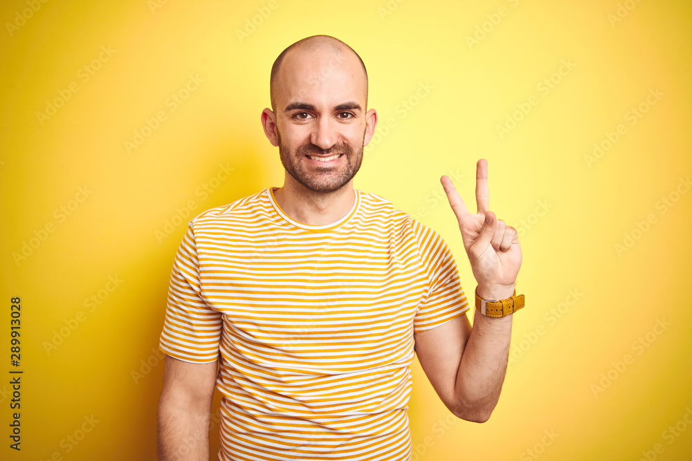Young bald man with beard wearing casual striped t-shirt over yellow isolated background showing and pointing up with fingers number two while smiling confident and happy. Young bald man with beard wearing casual striped t-shirt over yellow isolated background showing and pointing up with fingers number two while smiling confident and happy.