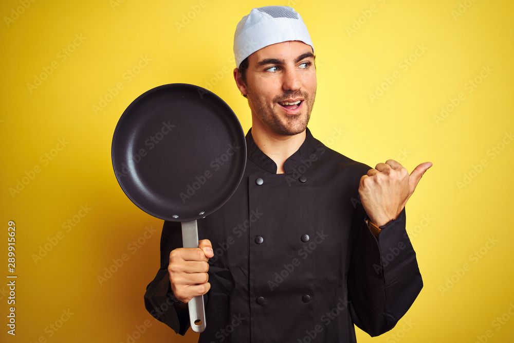 Young chef man wearing uniform and hat holding cook pan over isolated yellow background pointing and showing with thumb up to the side with happy face smiling Young chef man wearing uniform and hat holding cook pan over isolated yellow background pointing and showing with thumb up to the side with happy face smiling