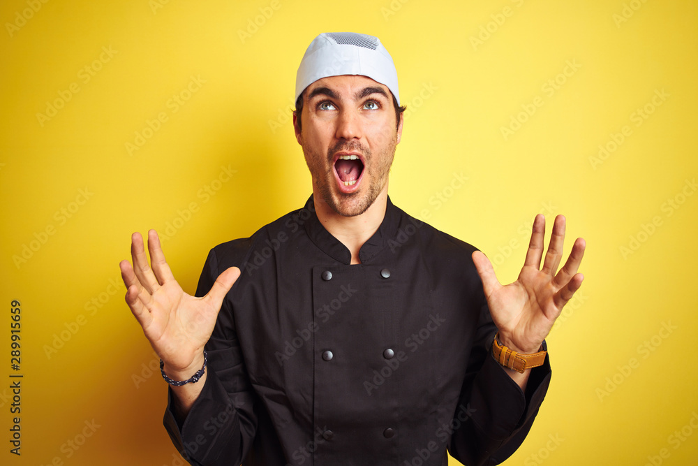 Young handsome chef man cooking wearing uniform and hat over isolated yellow background crazy and mad shouting and yelling with aggressive expression and arms raised. Frustration concept.